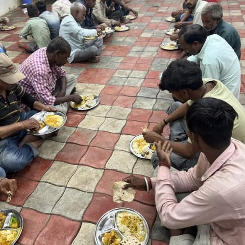 People sitting on the floor in rows eating a meal served on metal plates at a community food event.